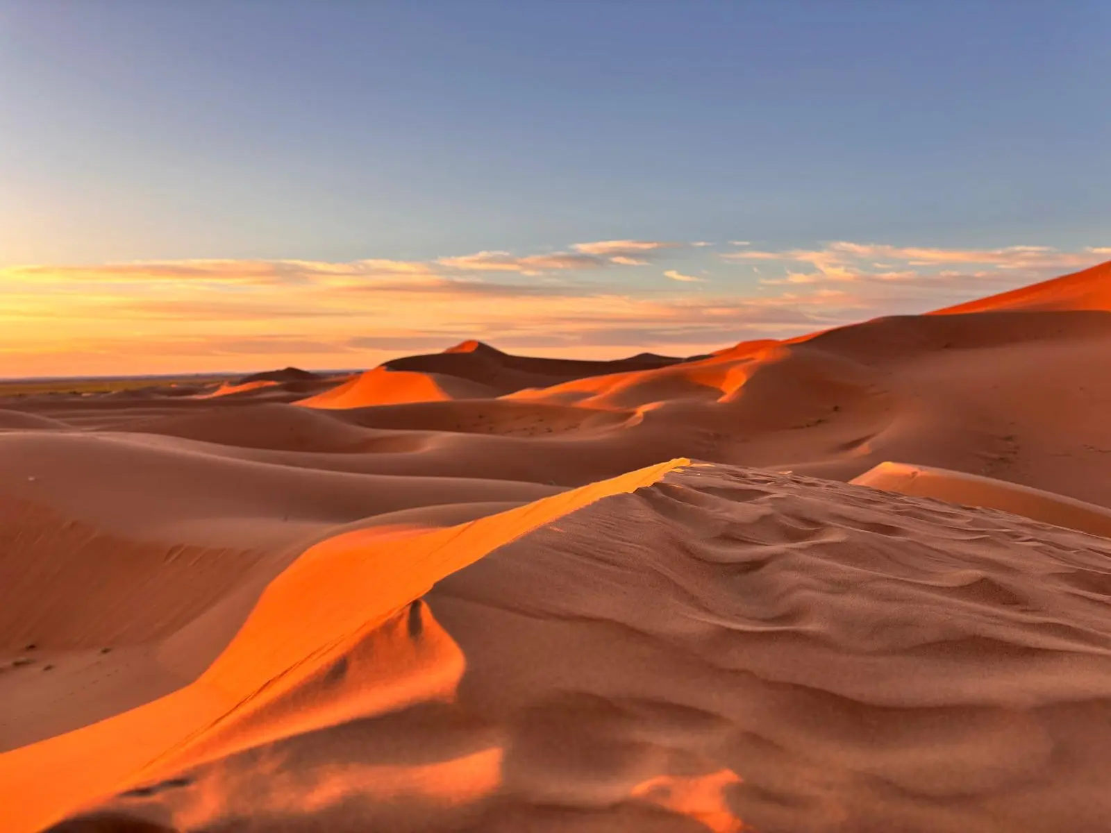 Vast sand dunes glowing orange and amber under a dramatic sunrise or sunset sky, casting long shadows across the desert.